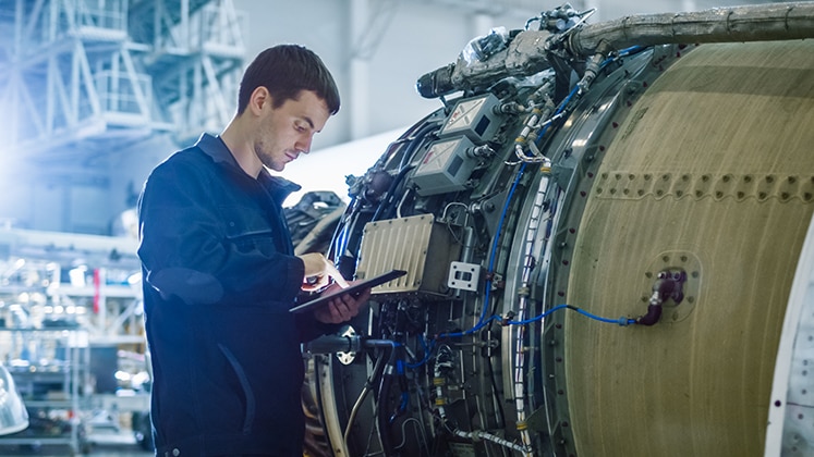 Aircraft maintenance technician works on an engine while referencing Maintenance Performance Toolbox on a tablet